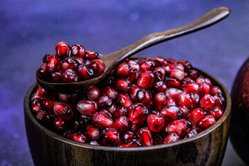 A full wooden bowl with pomegranate seeds on which is a wooden spoon. A bowl full of seeds with a spoonful of seeds on a blue background.