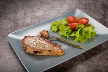 Roast pork loin, on a rectangular plate of gray color, with lettuce leaves, rosemary and tomatoes, on a gray background