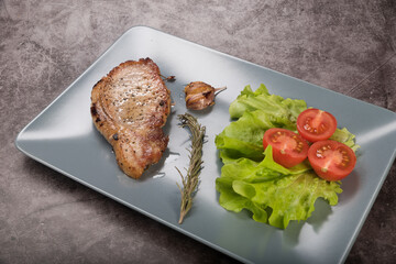 Roast pork loin, on a rectangular plate of gray color, with lettuce leaves, rosemary and tomatoes, on a gray background