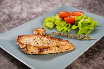 Roast pork loin, on a rectangular plate of gray color, with lettuce leaves, rosemary and tomatoes, on a gray background