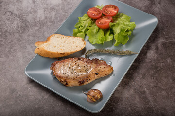 Roast pork loin, on a rectangular plate of gray color, with lettuce leaves, rosemary a piece of baguette and tomatoes, on a gray background