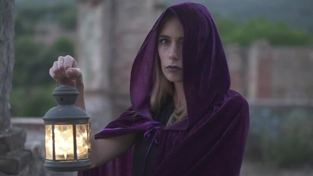 Mysterious young woman wearing a cloak and a hood covering her head staring at the camera and holding an old lantern. Role game female cosplayer