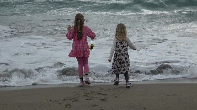 Two children are standing on a beach. A small wave approaches and the girls run away laughing not to get wet. The wave wash over their shoes and the younger girl stumbles and falls.