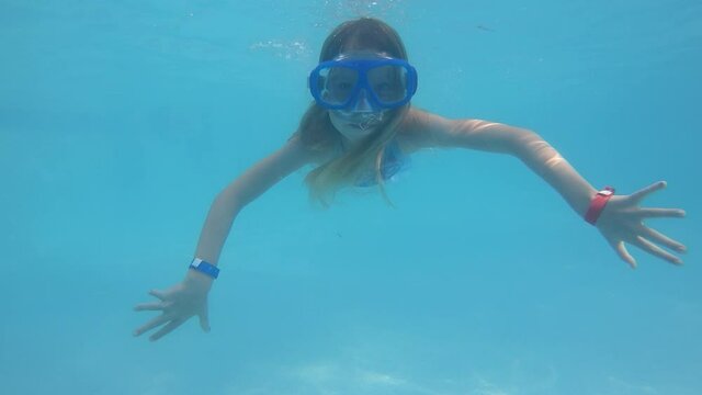 A younger girl dives and swims underwater towards the camera in a pool wearing a mask. The camera starts over the surface and dives to see the girl. Everything is filmed in slow motion.