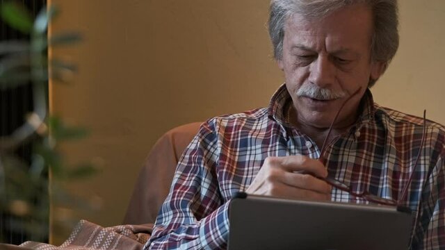 Front View Of A Man Sitting On Couch Using Tablet With Gray-Haired, Takes Off His Glasses And Looks In Front - Slow Motion Shot