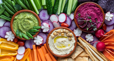 Top down close up view of three colourful hummus dips surrounded by various cut vegetables, pita...