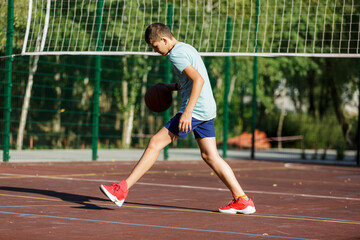 Fototapeta premium Cute young boy plays basketball on street playground in summer. Teenager in green t shirt with orange basketball ball outside. Hobby, active lifestyle, sport activity for kids.