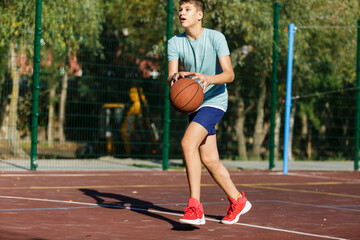 Fototapeta na wymiar Cute young boy plays basketball on street playground in summer. Teenager in green t shirt with orange basketball ball outside. Hobby, active lifestyle, sport activity for kids.