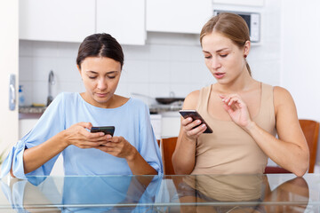 Two young girls having emotional conversation and looking at smartphone in kitchen