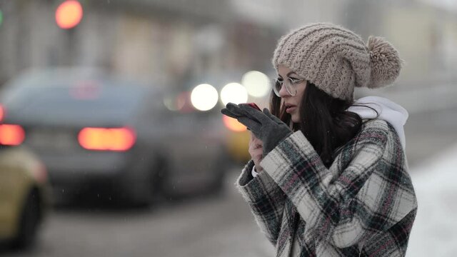 A beautiful young dark-haired woman in round glasses and a plaid coat walks around the city on a snowy winter day and talks on the phone.
