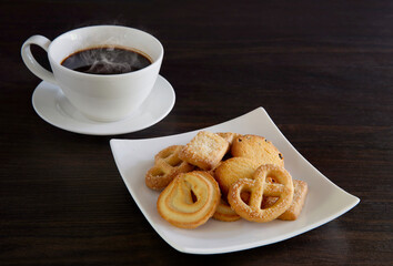 Butter cookies in white dish and black coffee in white cup all put on dark wooden background
