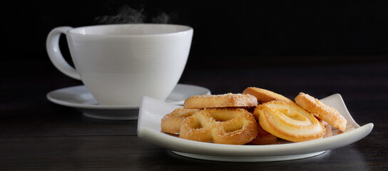 Butter cookies in white dish and black coffee in white cup all put on dark wooden background