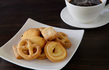 Butter cookies in white dish and black coffee in white cup all put on dark wooden background