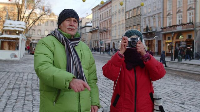 Senior wife and husband tourists taking photo pictures on retro camera, walking, gesturing on winter city street in Lviv, Ukraine. Family vacation activities and photography. Life after retirement