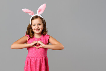 Happy easter. Cheerful little girl in a pink dress with polka dots on a gray background. A child in a rabbit costume shows a heart shape with his hands.