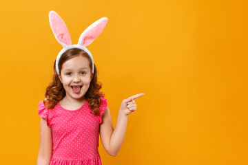 Cheerful happy cute little girl in Easter bunny ears on a yellow background. The child points with...