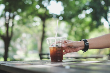 man hand touching a plastic cup of iced americano (black coffee) on the wood plank table in the garden