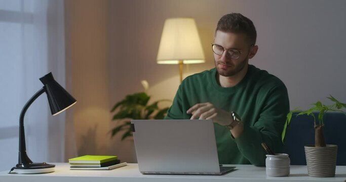 man with glasses is sitting in front of laptop, thinking and typing on keyboard, working at evening at home