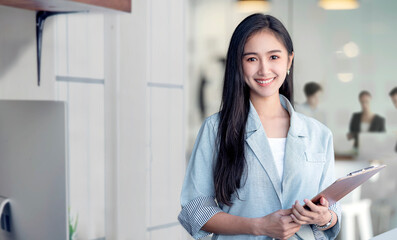 Portrait of young asian woman in casual wear holdng clipboard, smiling and looking at camera while standing in modern office room.