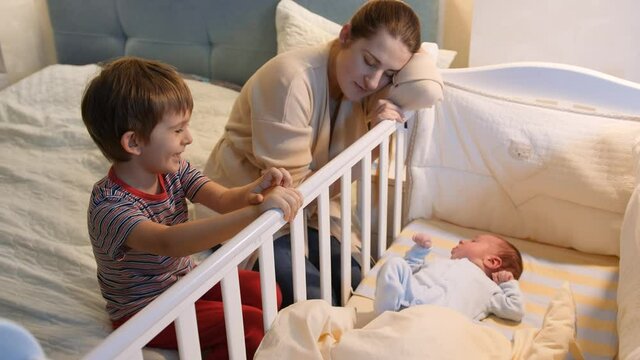 Older boy helping his tired mother rocking baby ccrib in bedroom at night. Children helping parents. Motherhood and sleepless nights