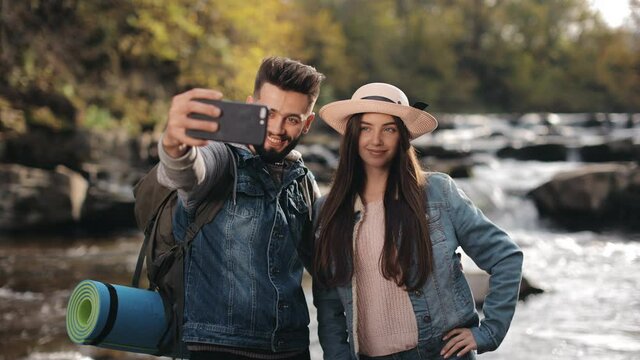 A young woman and a man are standing on the shore of a mountain river. The woman is adjusting her hat. They are taking selfies and smiling. 4K.