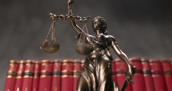 bronze blindfolded goddess representing law statue spinning, holding a scale and sword symbolizing impartiality and two broad sources of law in a university room with a background made of red books 