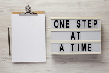 'One step at a time' on a lightbox, clipboard with blank sheet of paper on a white wooden...