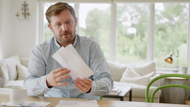 Smiling man sitting at table at home reviewing domestic finances opens letter containing good news - shot in slow motion
