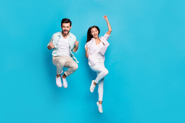 Photo of funny lucky young lovers dressed casual shirts jumping high rising fists isolated blue...