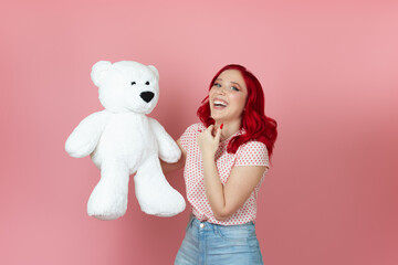 a smiling, delighted young woman with red hair holds a large white teddy bear isolated on a pink background.