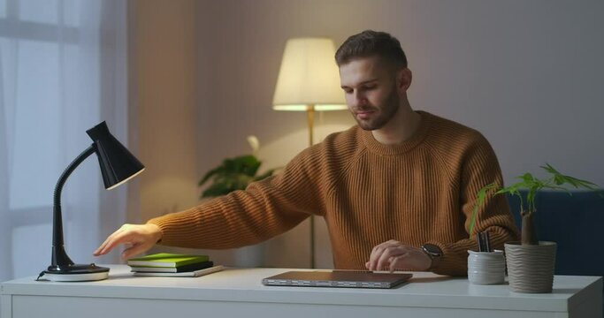 working at home, young man is opening laptop and switching on table lamp at evening, online remote work