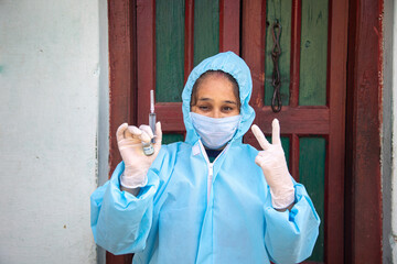 Young woman doctor in medical uniform holding a syringe and coronavirus vaccine, standing in front of village door.