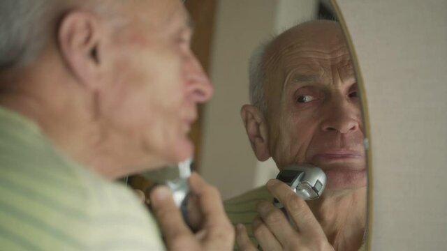 Serious older man looking in his reflection in mirror and shaving. Concept daily morning procedure.