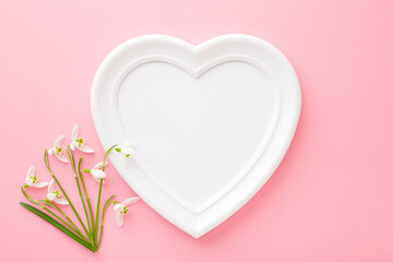 Frame of white heart shape and fresh snowdrops on light pink table. Pastel color. Love and happiness concept. Empty place for cute, emotional, sentimental text, quote or sayings. Closeup. Top view.