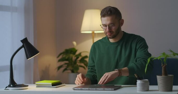 young man with glasses is using laptop at evening at home, opening notebook and switching on table lamp