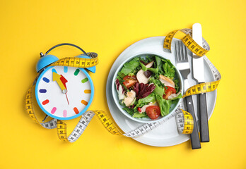 Plate of tasty salad, alarm clock and measuring tape on yellow background, flat lay. Nutrition regime