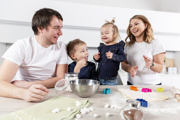 A young family with two small children in the kitchen are preparing cookie dough and laughing. The pleasure of spending time together. Love and tenderness.