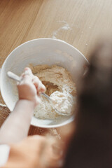 Hands of children stir a dough in the white bowl with spoon, on the wooden table in the kitchen. The process of mixing ingredients for pastry. Closeup, selective focus.