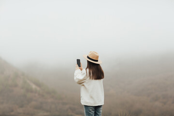 Girl on a viewpoint is taking a photo of the foggy mountain. 