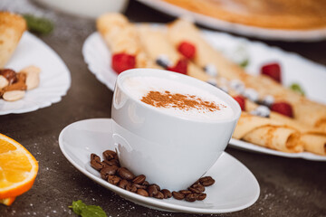 Cup of cappuccino coffee with coffee beans. In the background served pancakes with fruit