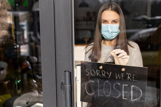 View through glass door female wearing medical mask and an apron holds closed sign board. Small business closed during coronavirus quarantine, lockdown