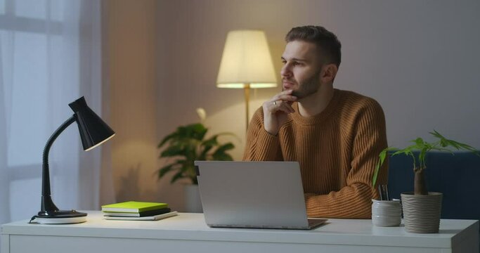 young guy is wearing brown sweater is sitting in front of table with laptop at evening, working at home