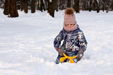 Little girl in purple overalls playing in a snowdrift with a snowball in a winter park outdoors