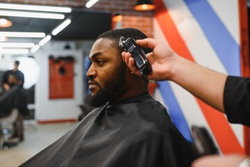 Portrait of young black man being trimmed with professional electric clipper machine in barbershop.Male beauty treatment concept. Young African guy getting new haircut in barber salon