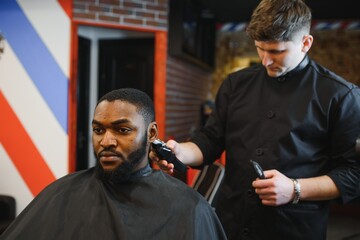 Portrait of young black man being trimmed with professional electric clipper machine in barbershop.Male beauty treatment concept. Young African guy getting new haircut in barber salon