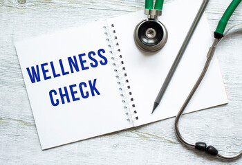 WELLNESS CHECK is written in a notebook on a wooden table next to stethoscope.