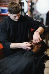 Side view of serious man with stylish modern haircut looking forward in barber shop. Hand of barber keeping straight razor and cutting trendy stripes on head of client.