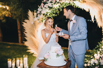 The groom in a gray suit feeds the blonde bride with a spoon with cake on the background of the...