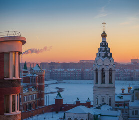 Fototapeta premium Winter frosty morning in Yoshkar-Ola city. Orange sunrise over the city. .Sunrise top view to the bell tower of the church and Kokshaga river embankment.