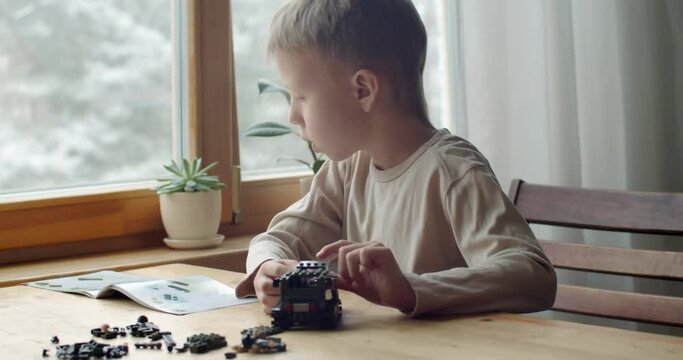 Young talented boy sitting at table engineering car at home by window slow motion. Concentrated child playing constructing bricks alone close up. Modern technologies activity on quarantine 